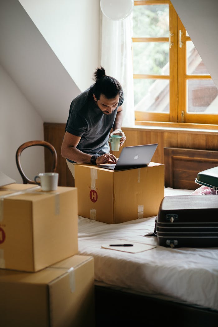 Ethnic male in casual clothes surfing internet while standing with cup of drink near stack of cardboard boxes and suitcase preparing to move into new house